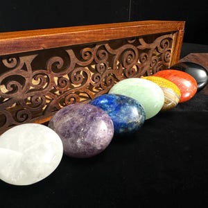 May include: A set of seven polished chakra stones in various colors, including white, purple, blue, green, yellow, orange, and black. The stones are displayed in front of a wooden box with a decorative carved design.