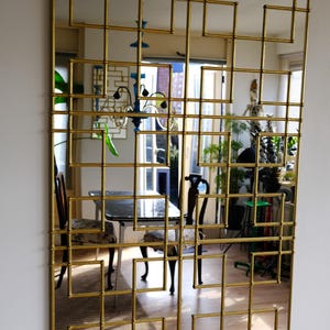 Pode incluir: Um grande espelho dourado com um design geométrico. O espelho é feito de vários quadrados e retângulos, criando um padrão de grade. O espelho reflete uma sala com móveis e plantas.