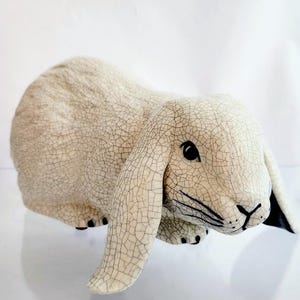 May include: A ceramic rabbit figurine in a cream color with a crackle glaze finish. The rabbit has long, floppy ears, black eyes, and black markings on its nose and feet. The figurine is sitting on a clear surface.