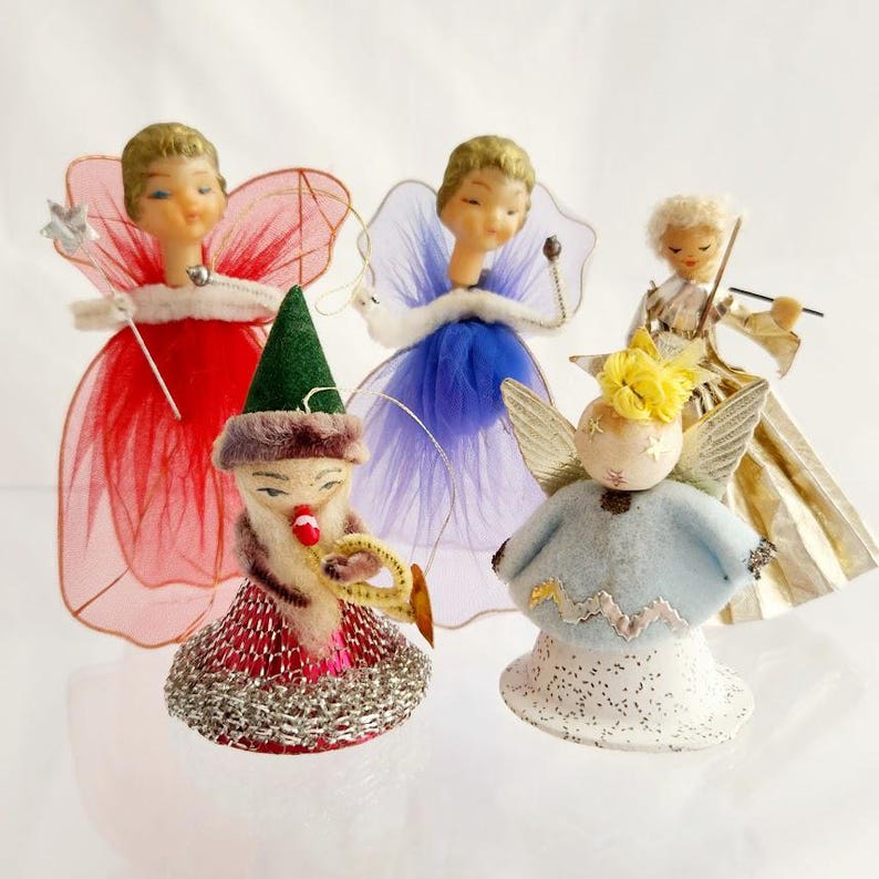 May include: A collection of vintage Christmas angel ornaments. The angels have delicate faces, fabric wings, and wear dresses in red, blue, gold, and light blue. One angel is playing a trumpet and wearing a green hat.