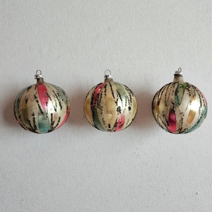 May include: Three spherical glass ornaments featuring a metallic gold finish with vertical stripes in pink and green. Each ornament is decorated with a textured black glitter pattern and topped with a small silver metal cap for hanging purposes.