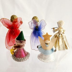 May include: A collection of vintage angel ornaments. The angels feature gold-colored hair, with wings made of red, blue, and sheer fabric. One angel plays a trumpet, while another holds a star. The ornaments are on a white surface.