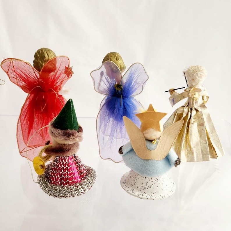 May include: A collection of vintage angel ornaments. The angels feature gold-colored hair, with wings made of red, blue, and sheer fabric. One angel plays a trumpet, while another holds a star. The ornaments are on a white surface.