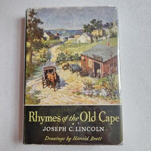 May include: A hardcover book titled Rhymes of the Old Cape by Joseph C. Lincoln with drawings by Harold Brett. The dust jacket features a colorful illustration of a horse-drawn carriage on a dirt road near a red barn and coastal village.