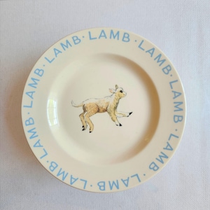 May include: A cream-colored ceramic plate featuring a central illustration of a small, light brown lamb in a running pose. The rim of the plate is decorated with the word LAMB printed repeatedly in a light blue sans-serif font around the edge.