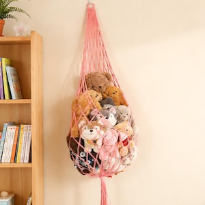 May include: A pink macrame hanging storage net filled with various stuffed animals. The net is suspended from a hook and is filled with teddy bears, dogs, and other plush toys. The net is a light pink color and has a tassel at the bottom.