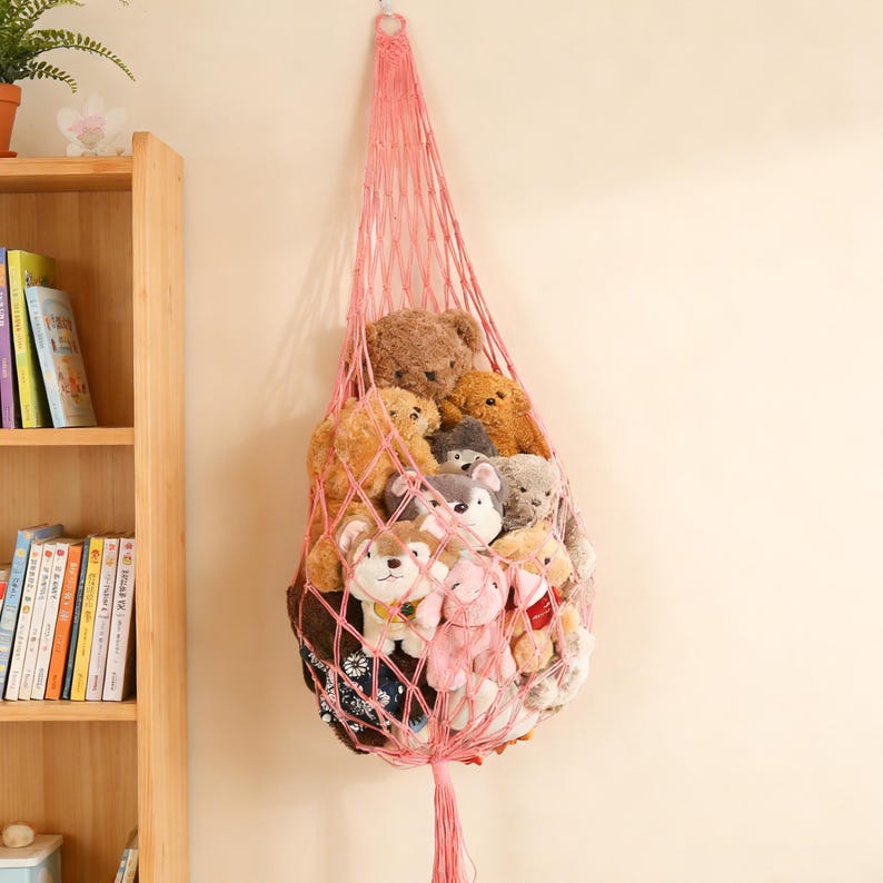May include: A pink macrame hanging storage net filled with various stuffed animals. The net is suspended from a hook and is filled with teddy bears, dogs, and other plush toys. The net is a light pink color and has a tassel at the bottom.