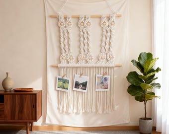 Könnte beinhalten: Ein cremefarbener Makramee-Fotorahmen mit drei angebrachten Fotos. Der Wandbehang besteht aus Seil und Holzdübeln. Die Fotos werden mit kleinen Holzklammern befestigt. Ein Holzschrank und eine Topfpflanze befinden sich im Hintergrund.