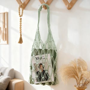 以下が含まれることがあります： 木製の壁掛けフックに吊るされた、薄緑色のマクラメバッグ。バッグには雑誌が入っており、表紙には「MAG」の文字が見えます。木製のビーズガーランドと、ドライフラワーが入った装飾的な花瓶も画像にあります。