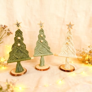 Puede incluir: Tres árboles de Navidad de macramé en tonos verdes y crema. Cada árbol tiene una estrella de madera en la parte superior y detalles de cuentas de madera. Los árboles están montados sobre pequeñas bases de madera.