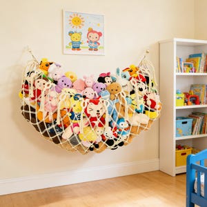 May include: A cream-colored, macrame-style toy hammock filled with various plush toys. The hammock is suspended from the wall, and a framed picture of cartoon characters hangs above it. A white bookshelf and a blue crib are visible in the background.