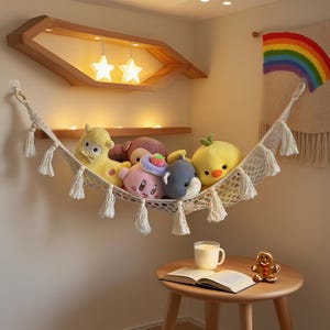 Könnte beinhalten: Eine cremefarbene Makramee-Hängematte, gefüllt mit Plüschtieren, hängt an einer Wand. Die Hängematte ist mit Quasten verziert. Ein Holzregal darüber hält sternförmige Lichter. Ein kleiner Holztisch enthält eine Tasse, ein offenes Buch und einen Lebkuchenmann.