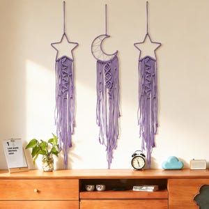 May include: Three lavender macrame wall hangings in star and crescent moon shapes. Each piece features intricate knotting and long, flowing fringe. The decor is displayed against a white wall, adding a touch of bohemian style to any space.