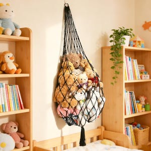 May include: A black macrame hanging storage net filled with various stuffed animals. The net is suspended from a hook on the wall and has a tassel at the bottom. The stuffed animals are of different sizes and colors, including teddy bears and other plush toys.