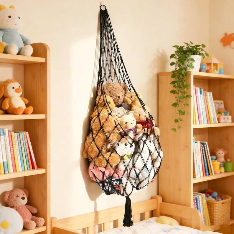 May include: A black macrame hanging storage net filled with various stuffed animals. The net is suspended from a hook on the wall and has a tassel at the bottom. The stuffed animals are of different sizes and colors, including teddy bears and other plush toys.