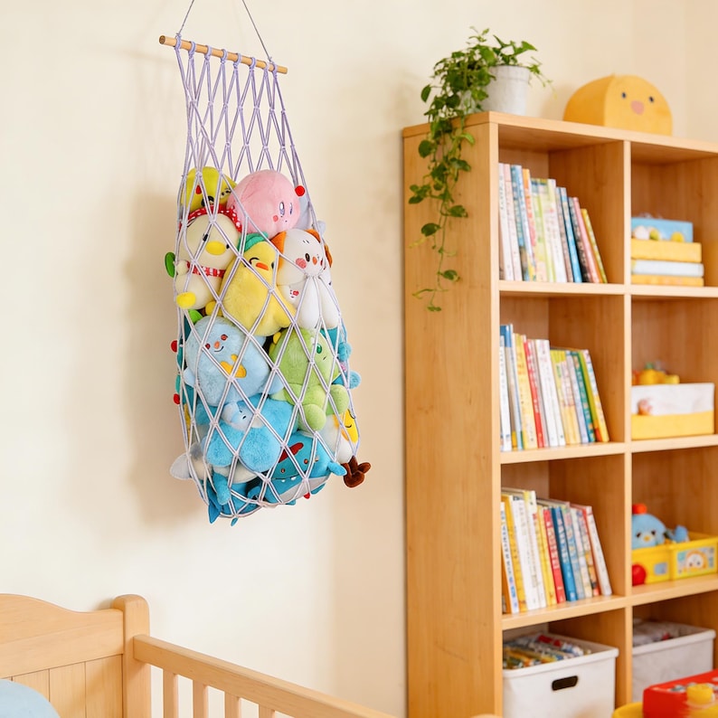 May include: A hanging, lavender-colored macrame net filled with colorful plush toys. The net is suspended from a wooden dowel and hangs against a cream-colored wall. The toys include various stuffed animals in shades of blue, yellow, pink, and green.