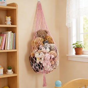 May include: A pink macrame hanging storage net filled with various stuffed animals. The net is suspended from a hook on a beige wall. The stuffed animals include teddy bears, dogs, and bunnies.
