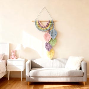 May include: A macrame wall hanging featuring a rainbow-colored semi-circle design with six pastel-colored leaf accents. The wall decor is suspended from a wooden dowel and hangs against a neutral wall. The leaves are pink, blue, green, purple, and yellow.