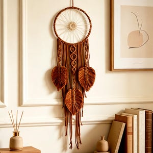 Può includere: Un acchiappasogni in macramè marrone con una cornice circolare e intricati dettagli intrecciati. Presenta tre accenti a forma di foglia e fili pendenti, creando un'estetica bohémien. L'arredamento della parete è di un tono caldo e terroso.