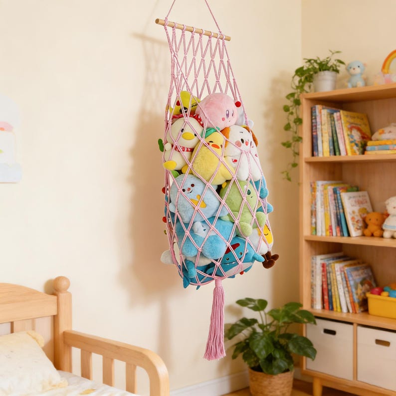 May include: A pink macrame hanging storage net filled with colorful plush toys. The net is suspended from a wooden dowel and features a long tassel. The toys include various animal shapes in shades of blue, yellow, green, and pink.