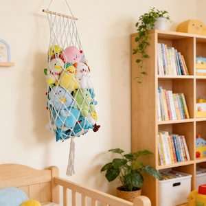 May include: A white macrame toy storage net filled with colorful plush toys, hanging on a wall. The net is suspended from a wooden dowel. The toys include various animal shapes in shades of blue, yellow, pink, and green.