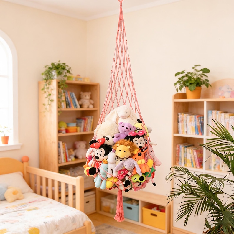 May include: A hanging pink net toy hammock filled with various stuffed animals. The hammock is suspended from the ceiling in a child's bedroom. The room also features wooden bookshelves, a bed, and a window.