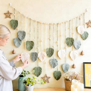 Könnte beinhalten: Ein Makramee-Blatt-Wandbehang in Grün-, Grau- und Cremetönen mit Holzperlen und Sternakzenten. Das dekorative Stück ist an einer Schnur aufgehängt und weist eine Vielzahl von Blattformen auf. Eine Glocke und ein Stern sind ebenfalls enthalten.