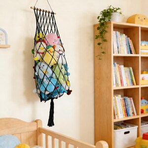 May include: A black macrame net toy storage bag filled with colorful plush toys, hanging on a wall. The bag is suspended from a wooden dowel and has a tassel at the bottom. The toys include various stuffed animals in shades of blue, yellow, pink, and green.
