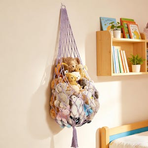 May include: A lavender macrame hanging toy storage net filled with various stuffed animals. The net is suspended from a hook on a white wall. The toys are a mix of colors and sizes, including teddy bears and other plush creatures.