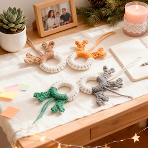 May include: Four handmade macrame Christmas ornaments in neutral colors. Each ornament features a circular design with a reindeer-like shape on top. The colors include beige, orange, green, and gray. They are displayed on a wooden surface with a succulent and a framed photo.