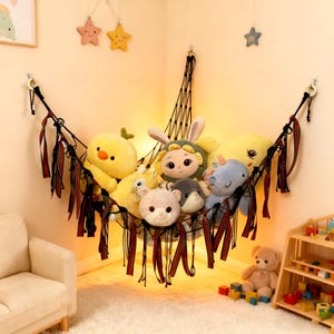 Può includere: Un'amaca a rete nera piena di vari peluche, tra cui un pulcino giallo, un coniglietto con la faccia a fiore e un unicorno blu. L'amaca è decorata con nastri rossi e neri ed è sospesa in un angolo di una stanza.