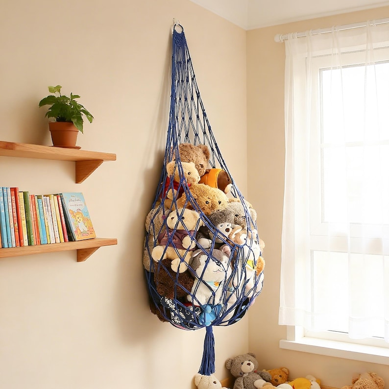 May include: A navy blue macrame toy hammock filled with various stuffed animals. The hammock is suspended from a wall hook, creating a space-saving storage solution. The room also features wooden shelves with books and a potted plant.