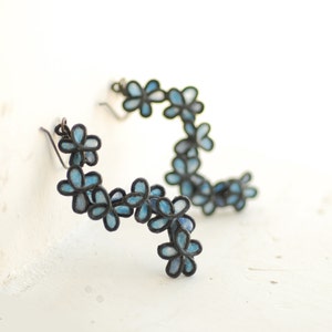 May include: A pair of floral earrings. Each earring features a series of small, light blue flowers with dark outlines. The flowers are arranged in a curved, cascading design. The earrings have a hook closure.