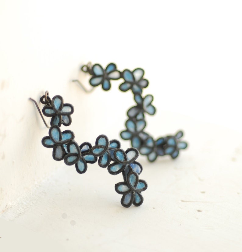 May include: A pair of floral earrings. Each earring features a series of small, light blue flowers with dark outlines. The flowers are arranged in a curved, cascading design. The earrings have a hook closure.