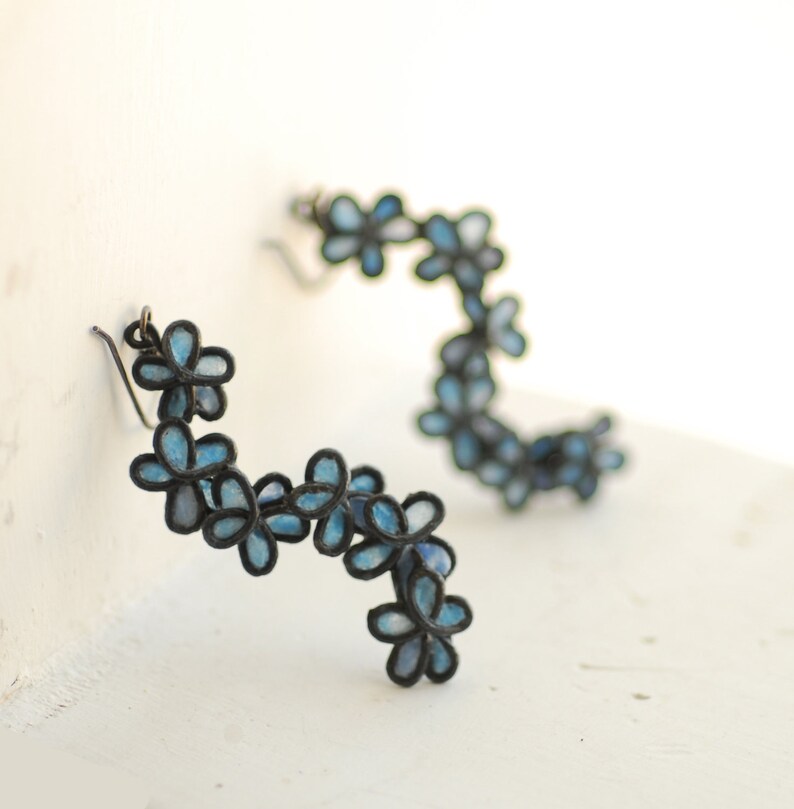 May include: A pair of floral earrings. Each earring features a curved design of small, blue flower shapes with black outlines. The earrings are set against a white background, highlighting the delicate details and color contrast.