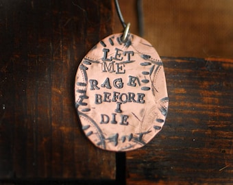 May include: A copper-colored pendant with the words "LET ME RAGE BEFORE I DIE" etched in black. The pendant is oval-shaped and has decorative markings around the edges. It hangs from a black cord.