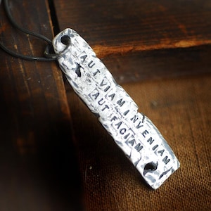 Puede incluir: Collar con colgante plateado con la frase latina "AUT VIAM INVENIAM AUT FACIAM". El colgante rectangular, de unos 5 cm de largo, tiene una superficie texturizada y está suspendido de un cordón negro.