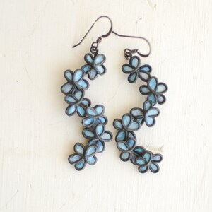 May include: A pair of dangle earrings featuring a floral design. Each earring has a series of light blue flower-shaped elements with dark gray outlines. The earrings hang from dark gray ear wires, creating a delicate and feminine accessory.
