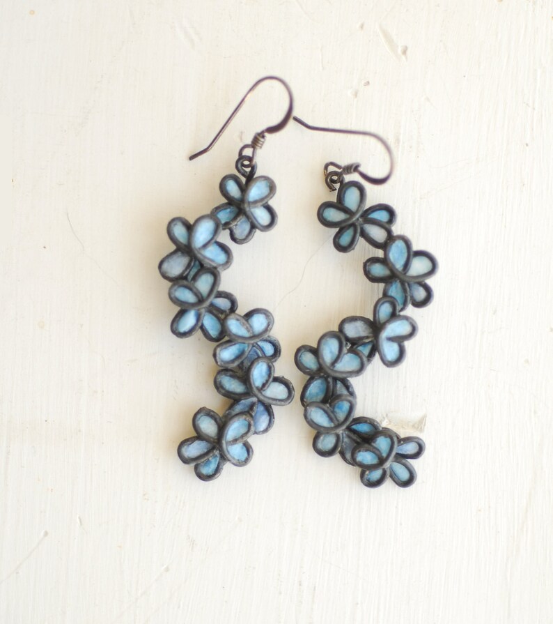 May include: A pair of dangle earrings featuring a floral design. Each earring has a series of light blue flower-shaped elements with dark gray outlines. The earrings hang from dark gray ear wires, creating a delicate and feminine accessory.