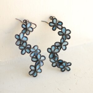 May include: A pair of floral earrings. Each earring features a series of light blue flower-shaped charms with dark outlines. The earrings have a curved design, creating a cascading effect. The metal has a dark, antique finish.