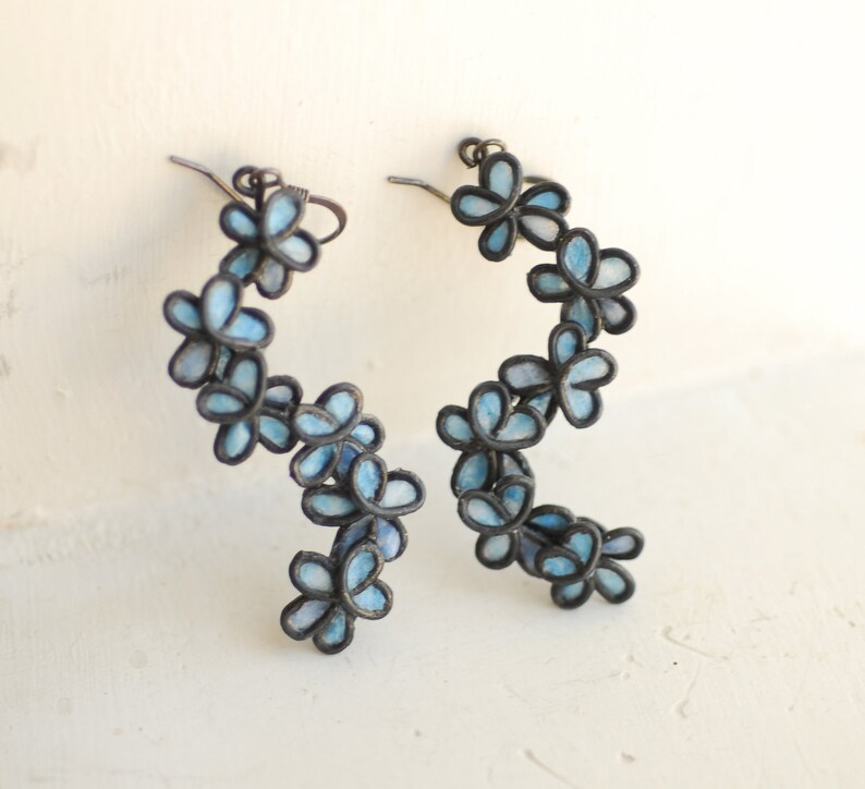 May include: A pair of floral earrings. Each earring features a series of light blue flower-shaped charms with dark outlines. The earrings have a curved design, creating a cascading effect. The metal has a dark, antique finish.