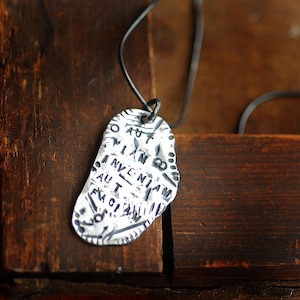 May include: A silver-colored pendant with a black cord. The pendant has an irregular shape and is inscribed with black text. The background is a dark wooden surface.