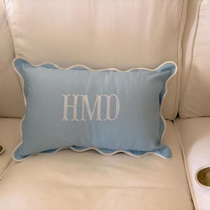 May include: A light blue rectangular pillow with white scalloped edges and white embroidered initials "HMD". The pillow is resting on a white sofa, with a green pillow visible on the left side.