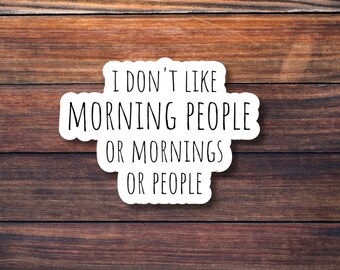 Op de afbeelding: Een witte sticker met zwarte tekst: "I DON'T LIKE MORNING PEOPLE OR MORNINGS OR PEOPLE" tegen een bruine houten achtergrond. De sticker heeft een afgeronde, onregelmatige vorm.