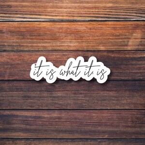 May include: White sticker with the text "it is what it is" on a brown wooden background.