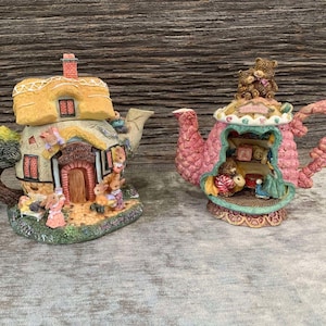 May include: Two ceramic teapots shaped like houses. One is a pink teapot with a teddy bear family inside. The other is a yellow teapot shaped like a cottage with the words "Bunny Tea House" on the front.