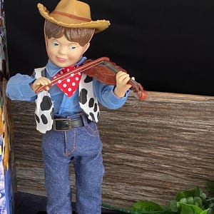 Könnte beinhalten: Figur eines Cowboys, der Geige spielt. Der Cowboy trägt einen braunen Hut, ein blaues Hemd, Jeans und eine Weste mit Kuhmuster. Der schwarze Sockel trägt den Schriftzug "COUNTRY COWBOY" in Goldbuchstaben.