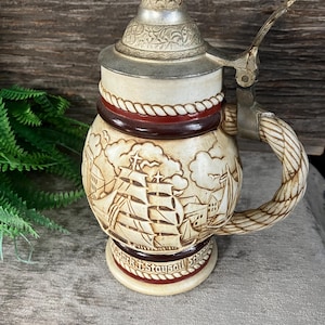 May include: A ceramic beer stein with a pewter lid and a rope-like handle. The stein features a detailed illustration of sailing ships and the text "Stark & Staysail" on the bottom.