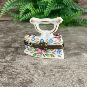 Op de afbeelding: Een wit porseleinen snuisterijendoosje in de vorm van een vintage strijkijzer, voorzien van handgeschilderde roze rozen, blauwe bloemen en gouden accenten. Het doosje heeft een messingkleurig metalen scharnier en een decoratieve sluiting.