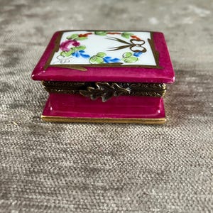 Op de afbeelding: Een klein rechthoekig porseleinen doosje met een fel magenta basis en deksel. De bovenkant heeft een wit inzetpaneel versierd met een geschilderde bloemenkrans in roze, blauw en groen, gecentreerd met een zwarte strik. Voorzien van een messing scharnier.